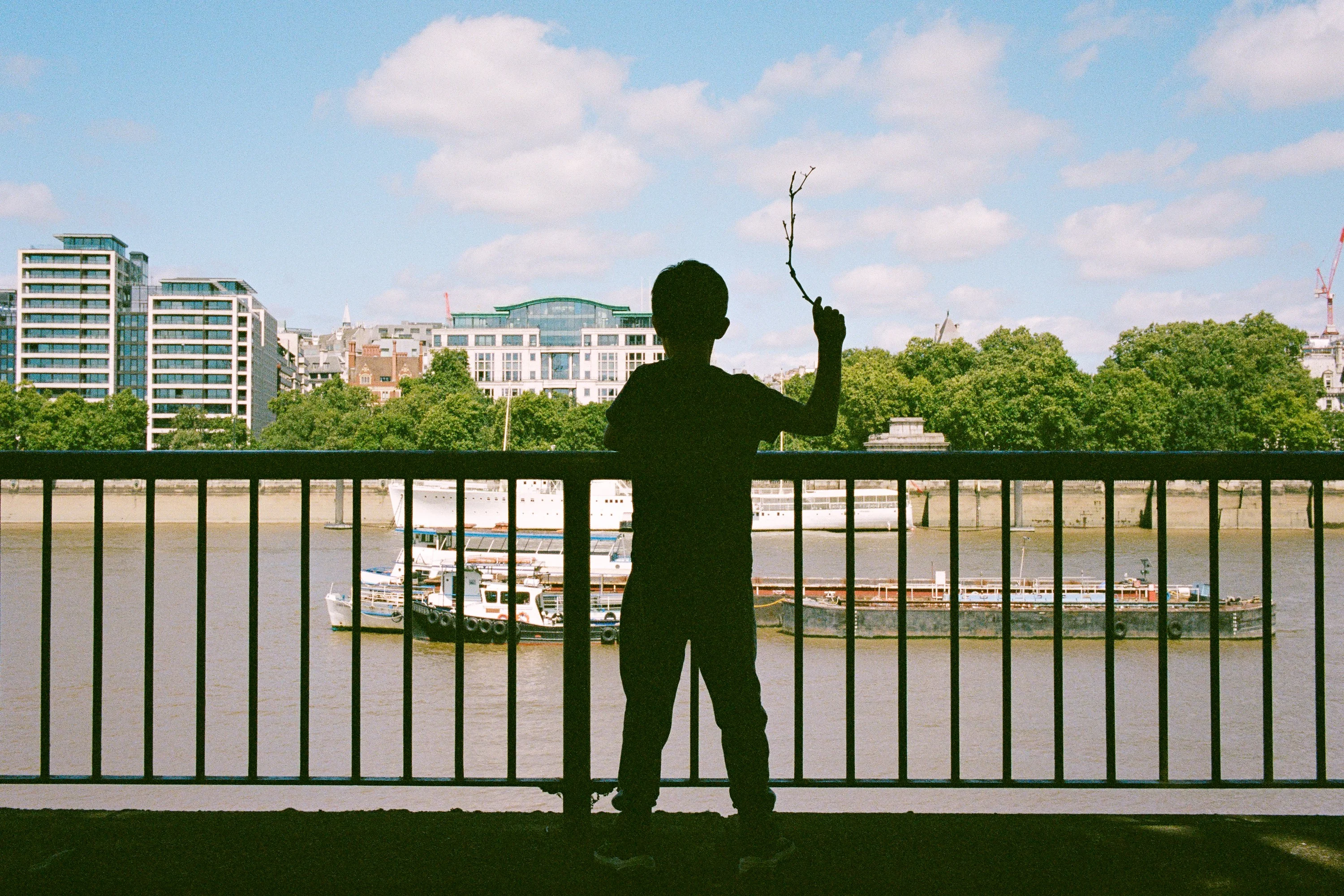 A boy in silhouette at the river railings holding up a stick
