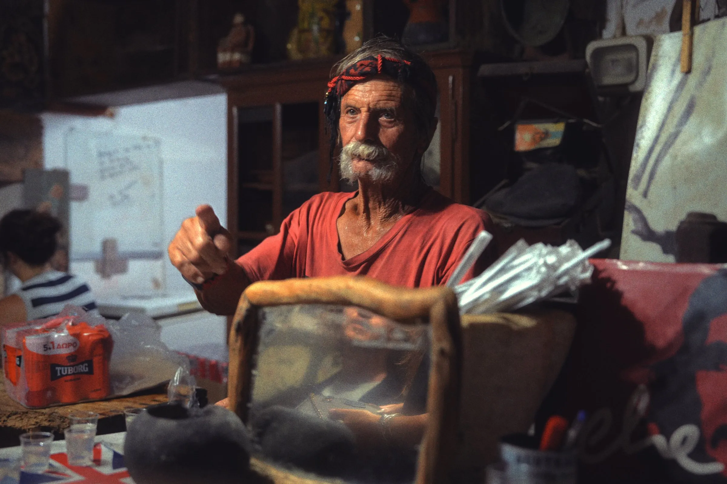 An old man with a white moustache and a red headband gestures from behind his cluttered bar