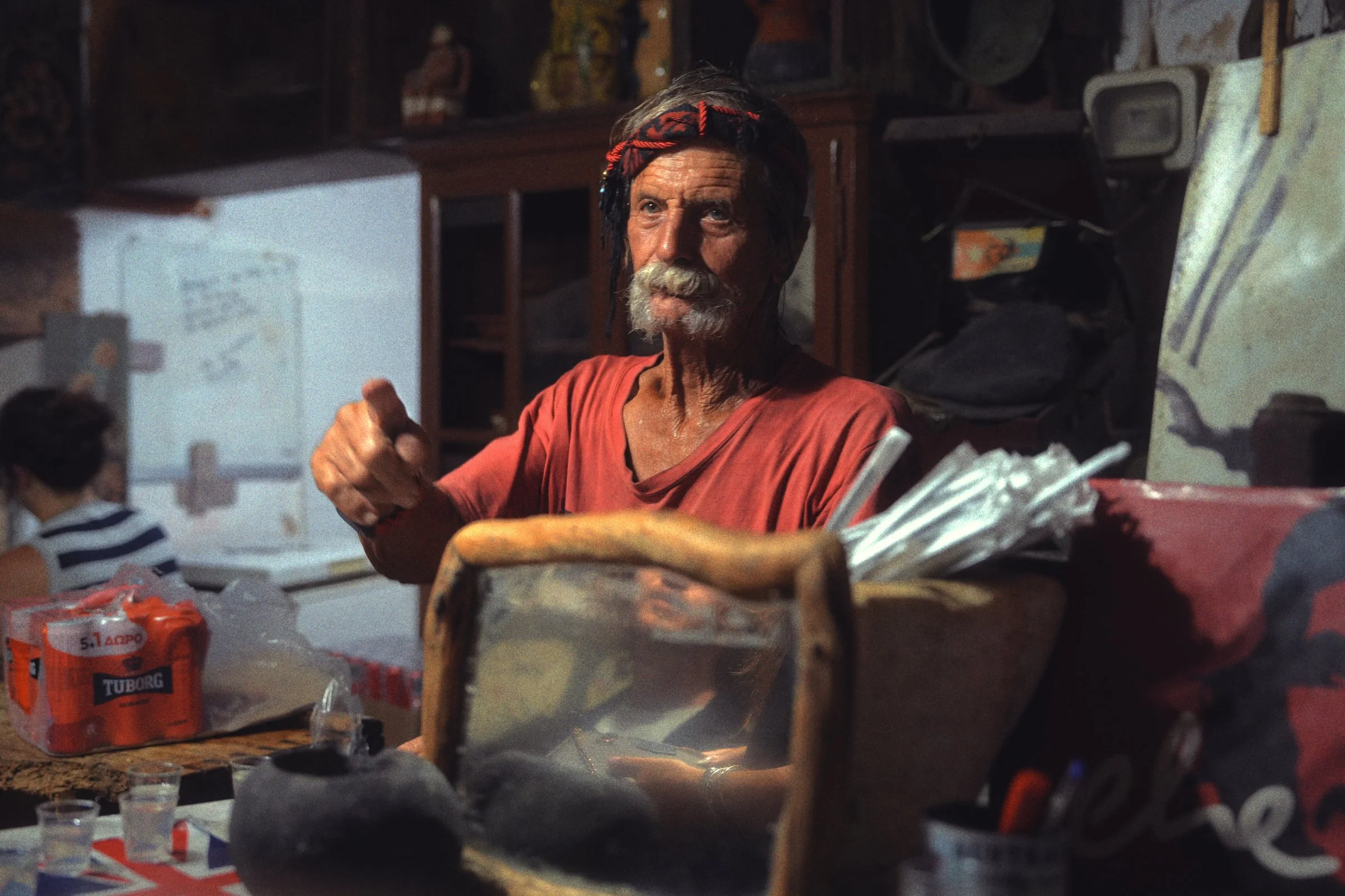 An old man with a white moustache and a red headband gestures from behind his cluttered bar