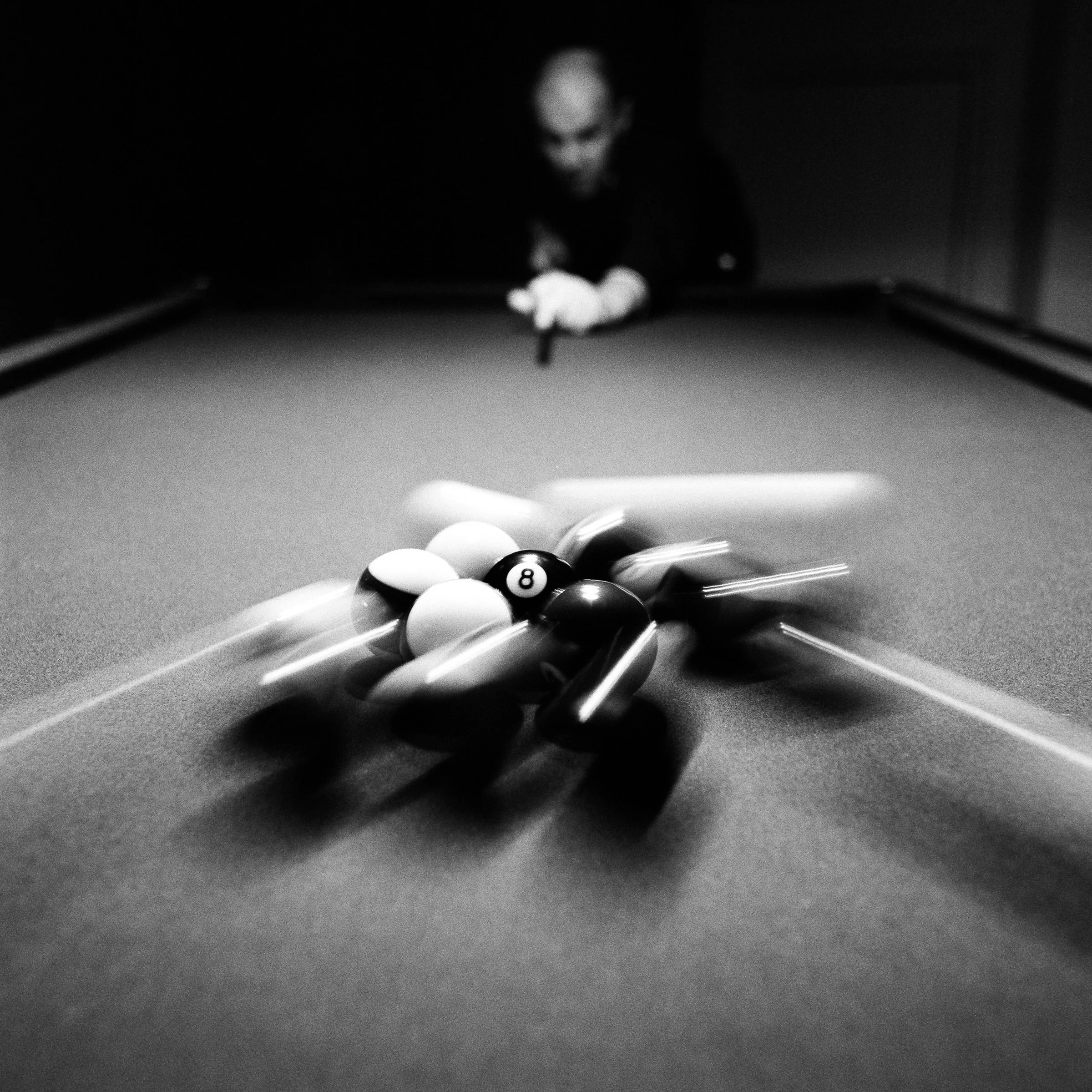 Black and white photo of a pool break, the balls blurring as the player watches from the dark