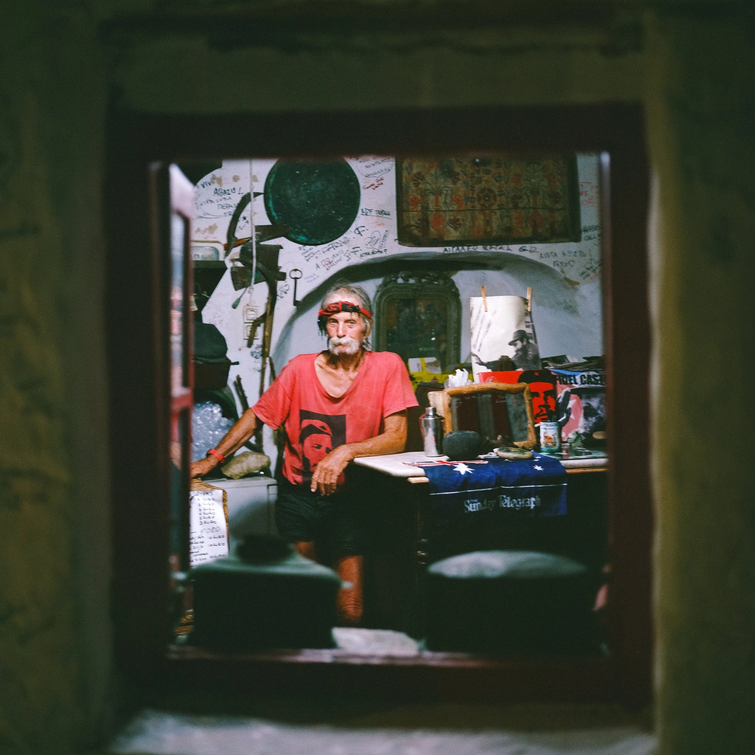 An old man in a red shirt framed by a doorway inside his cluttered shop