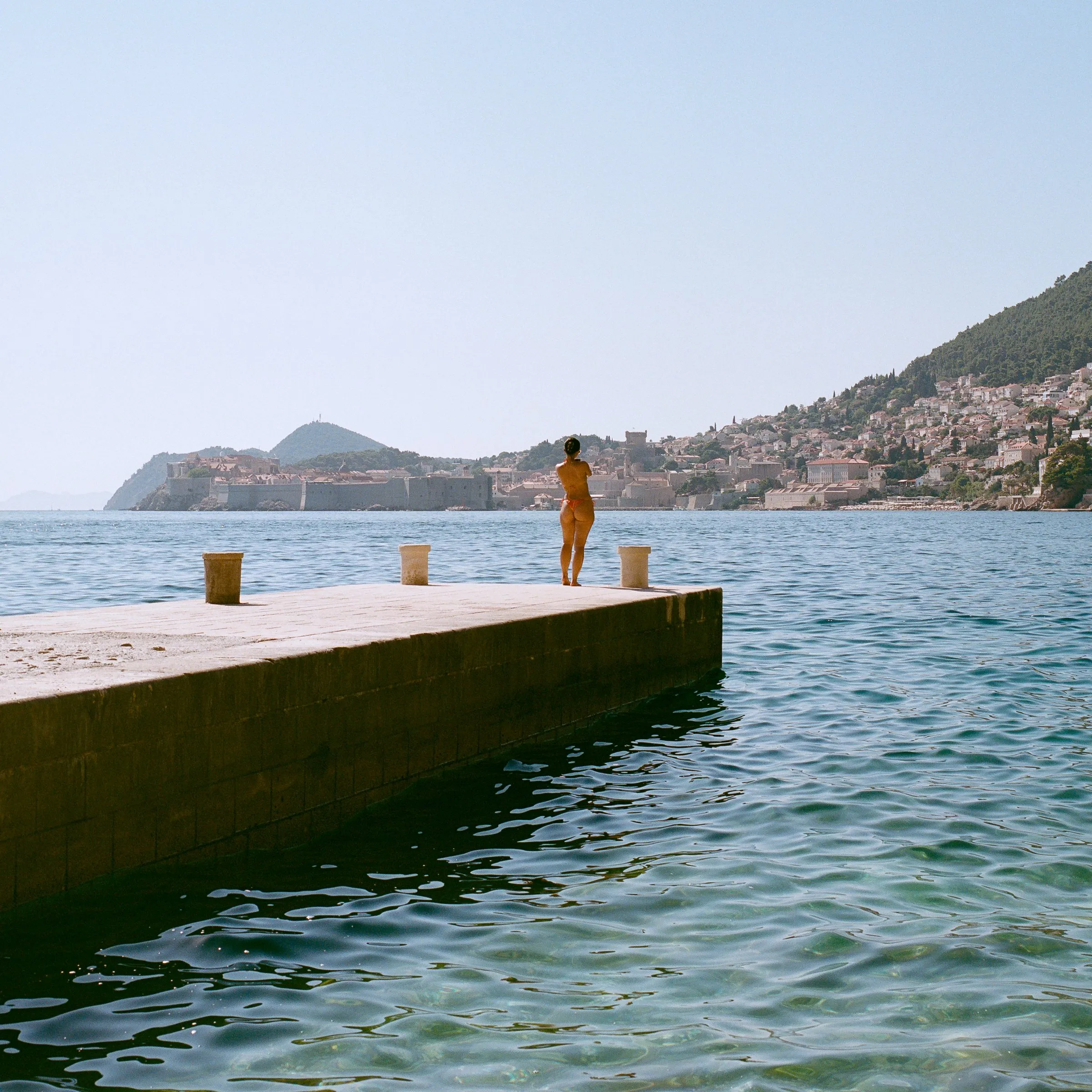 A swimmer stands at the end of a stone pier, with a hillside town across the water
