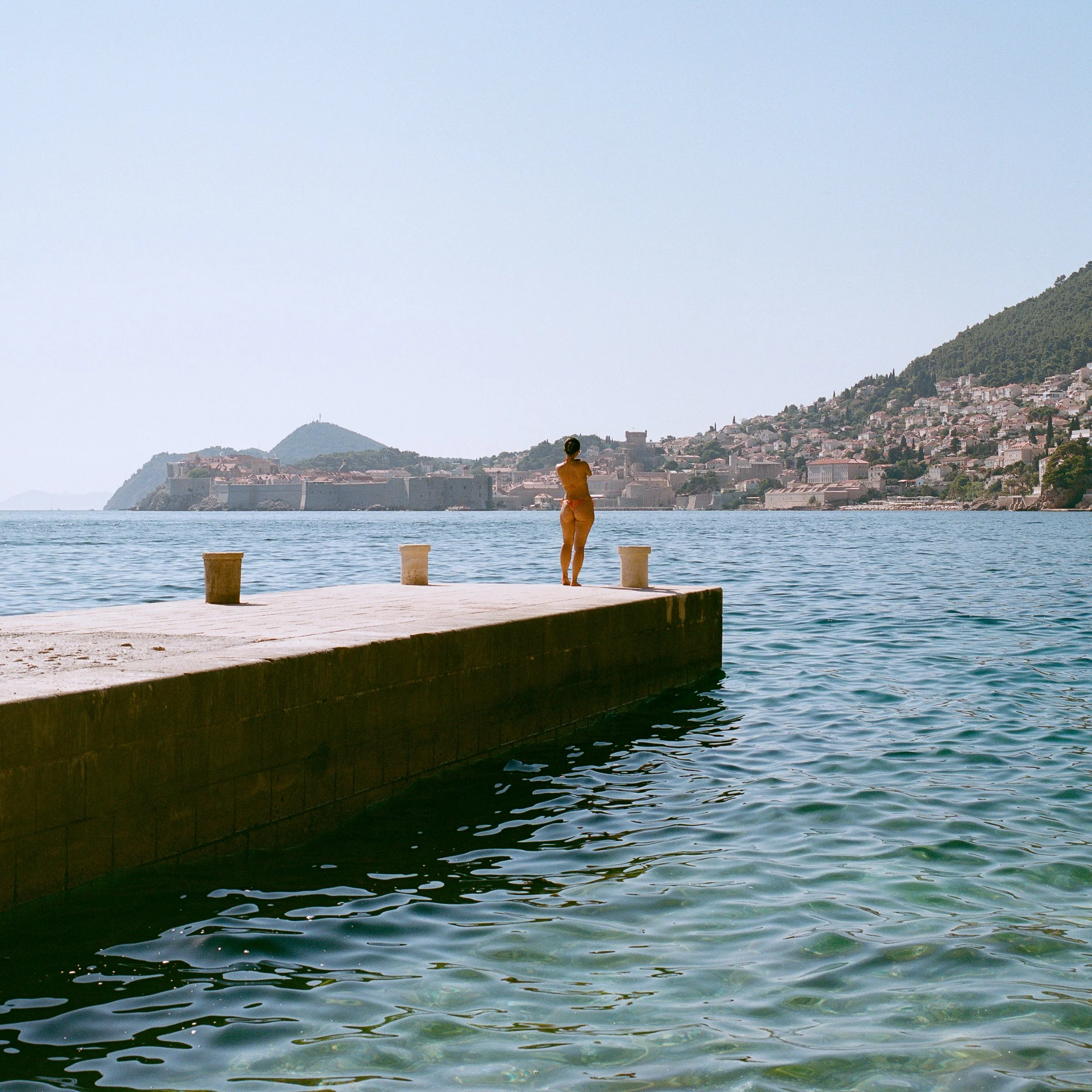 A swimmer stands at the end of a stone pier, with a hillside town across the water