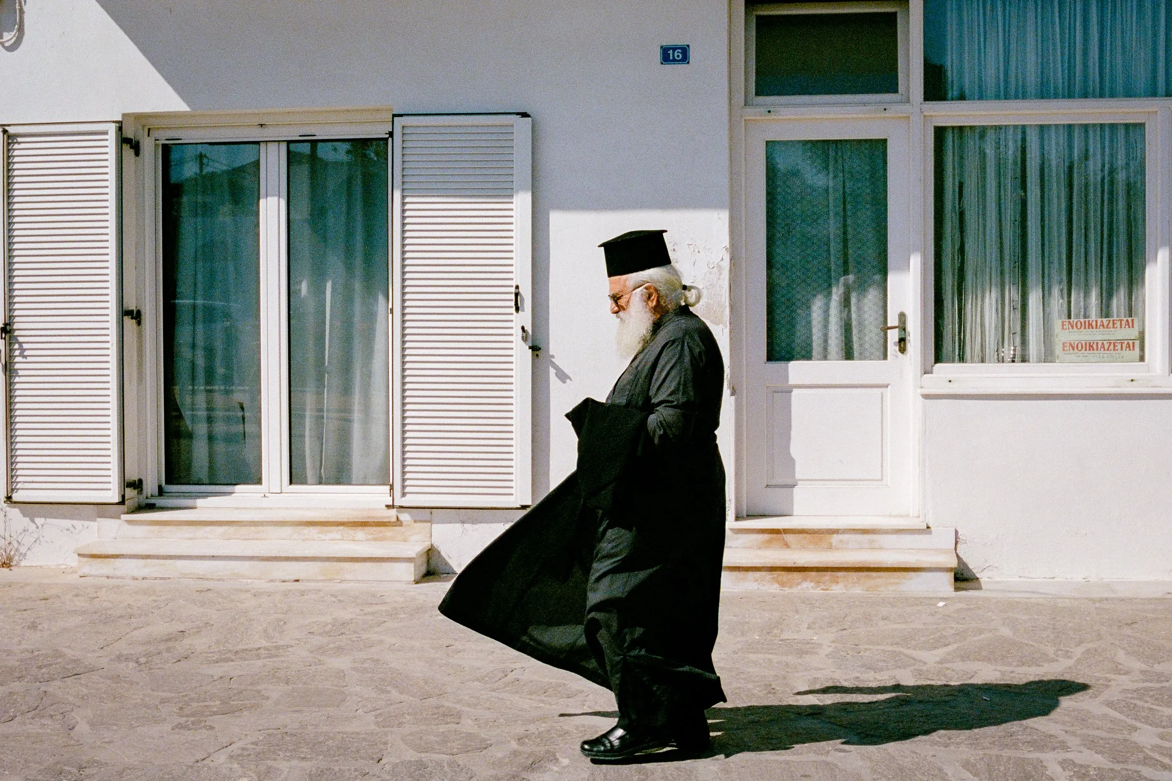 A priest in black robes walks across a white square, his robe lifted by the wind