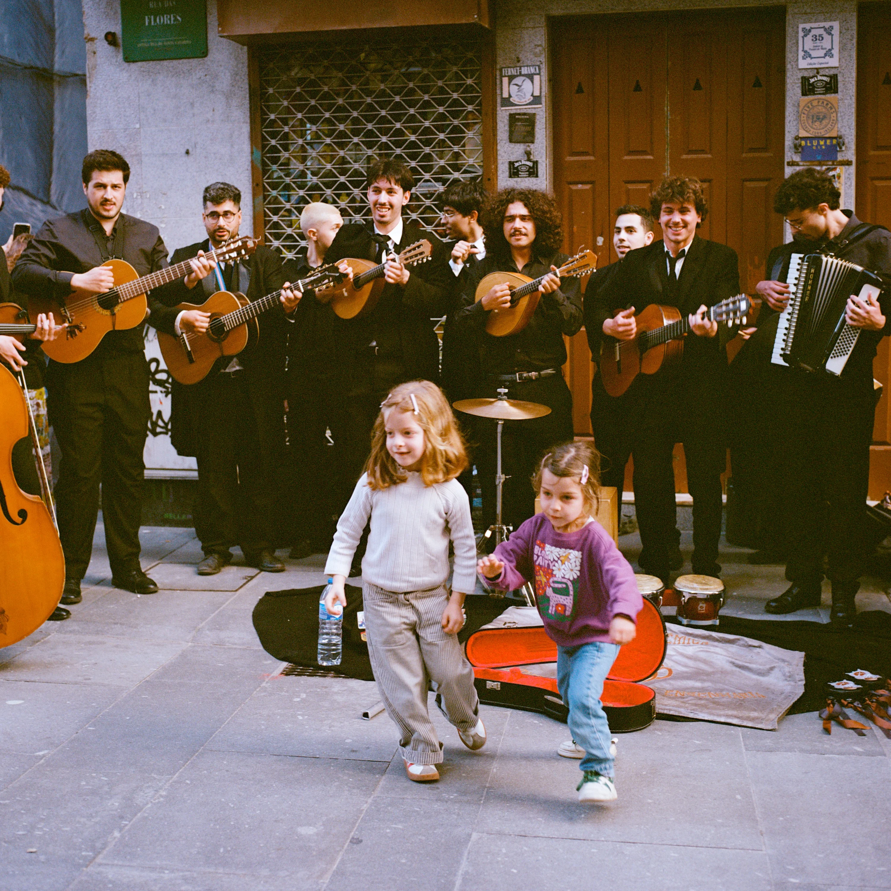 Two small children dance in front of a student music group in black capes