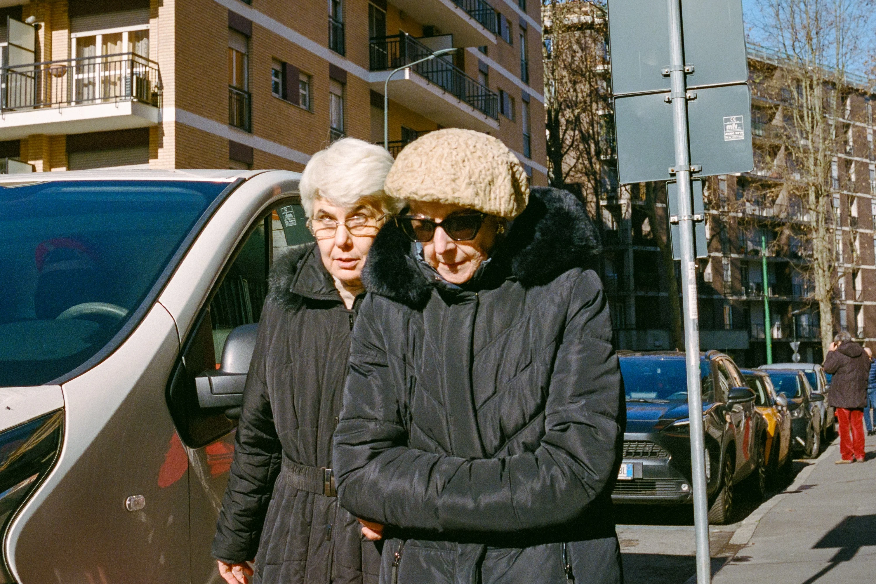 Two older women in winter coats walking past a van