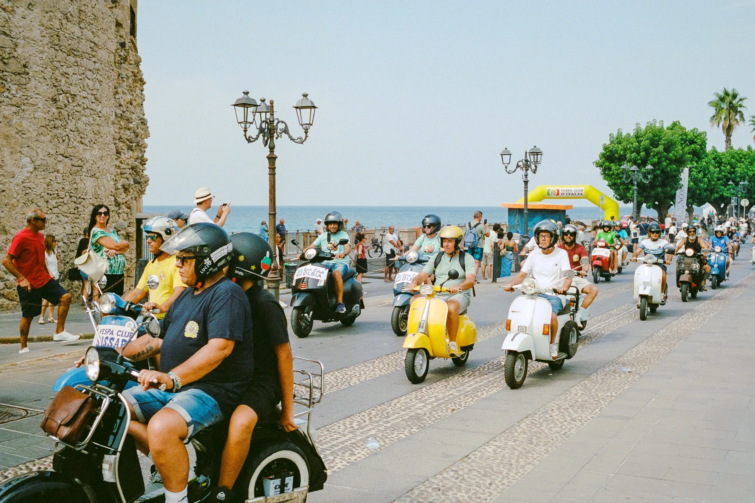 A pack of riders on vintage Vespas and scooters coming down a seafront road