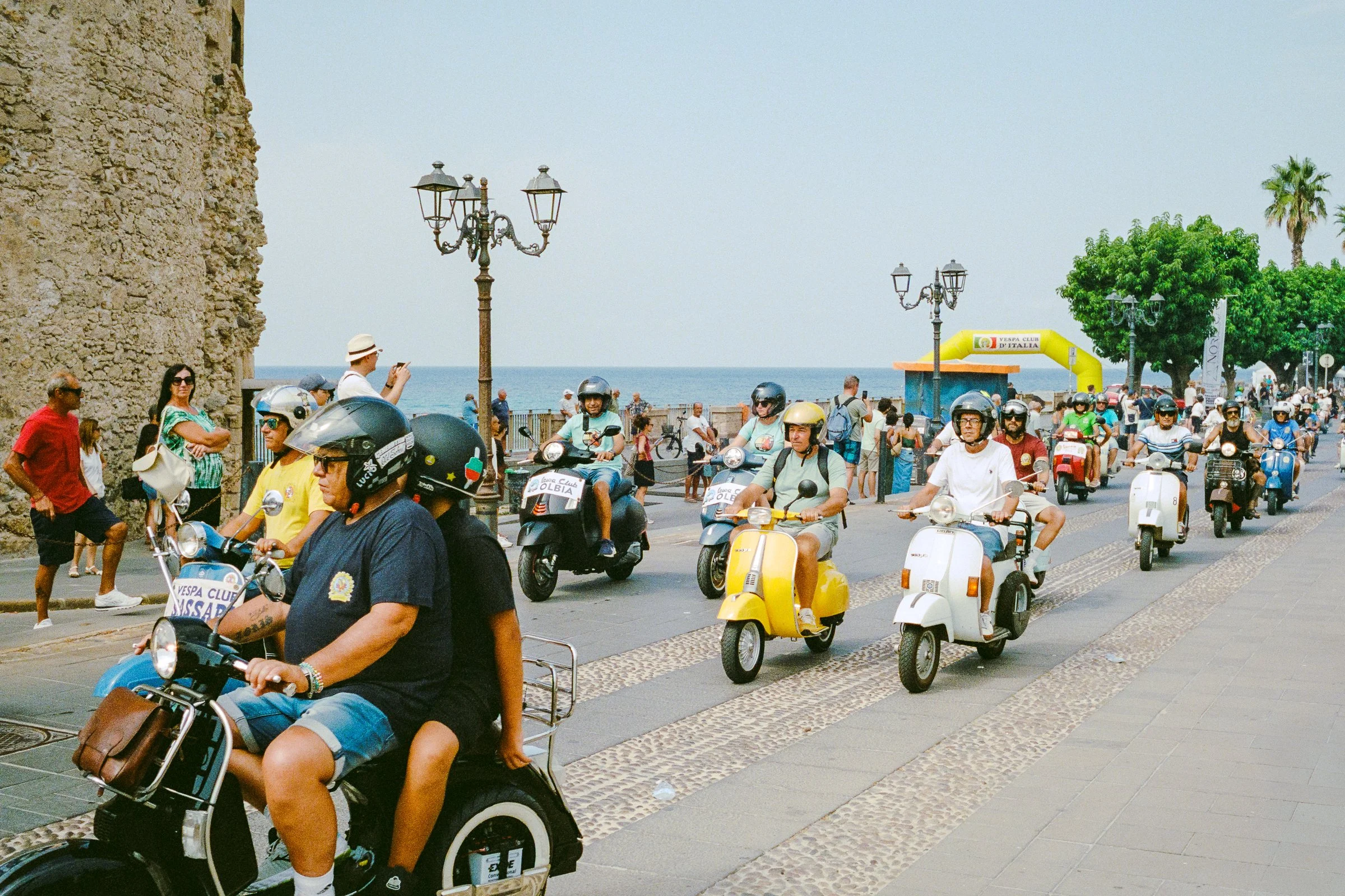 A pack of riders on vintage Vespas and scooters coming down a seafront road