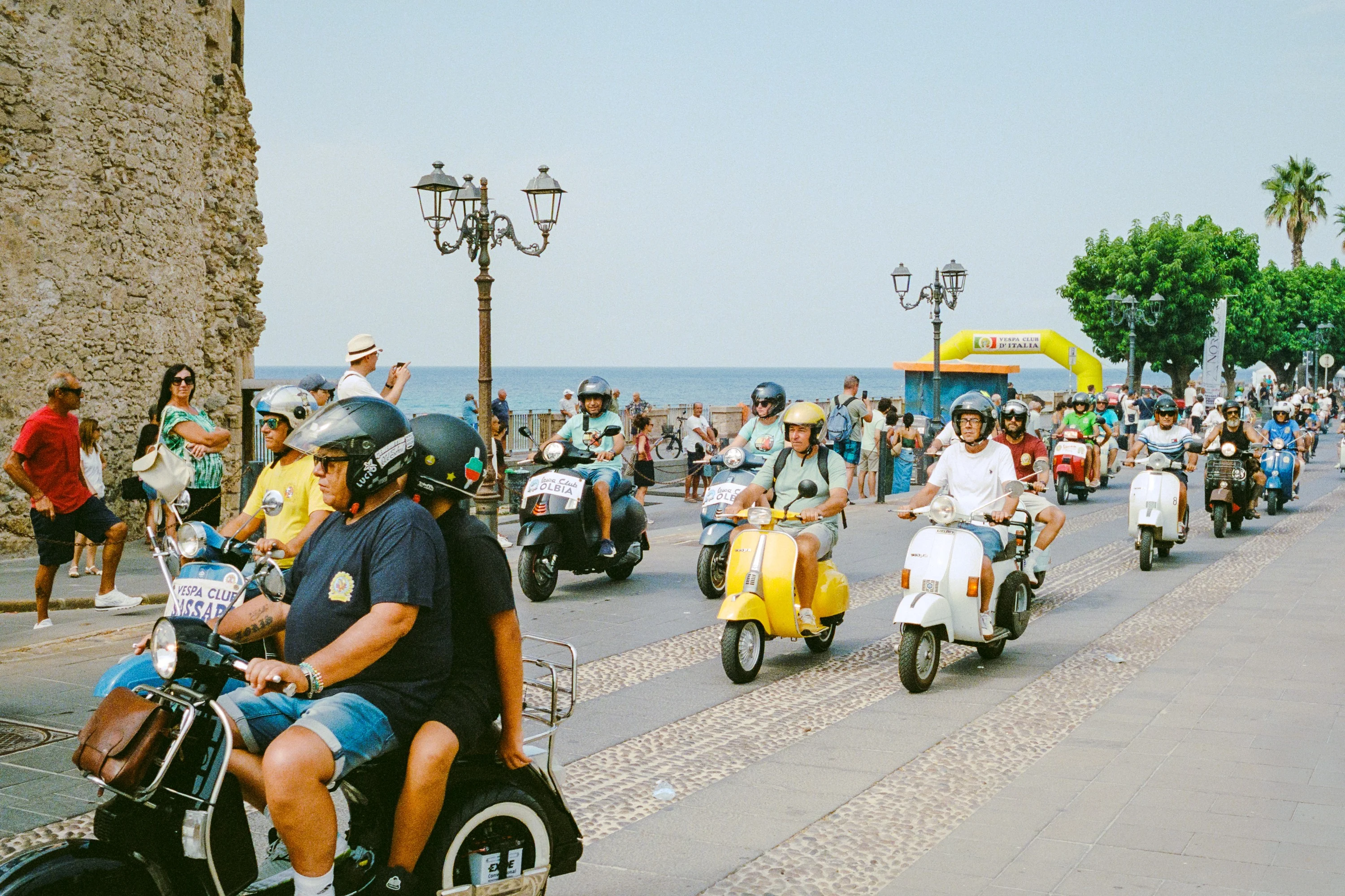 A pack of riders on vintage Vespas and scooters coming down a seafront road