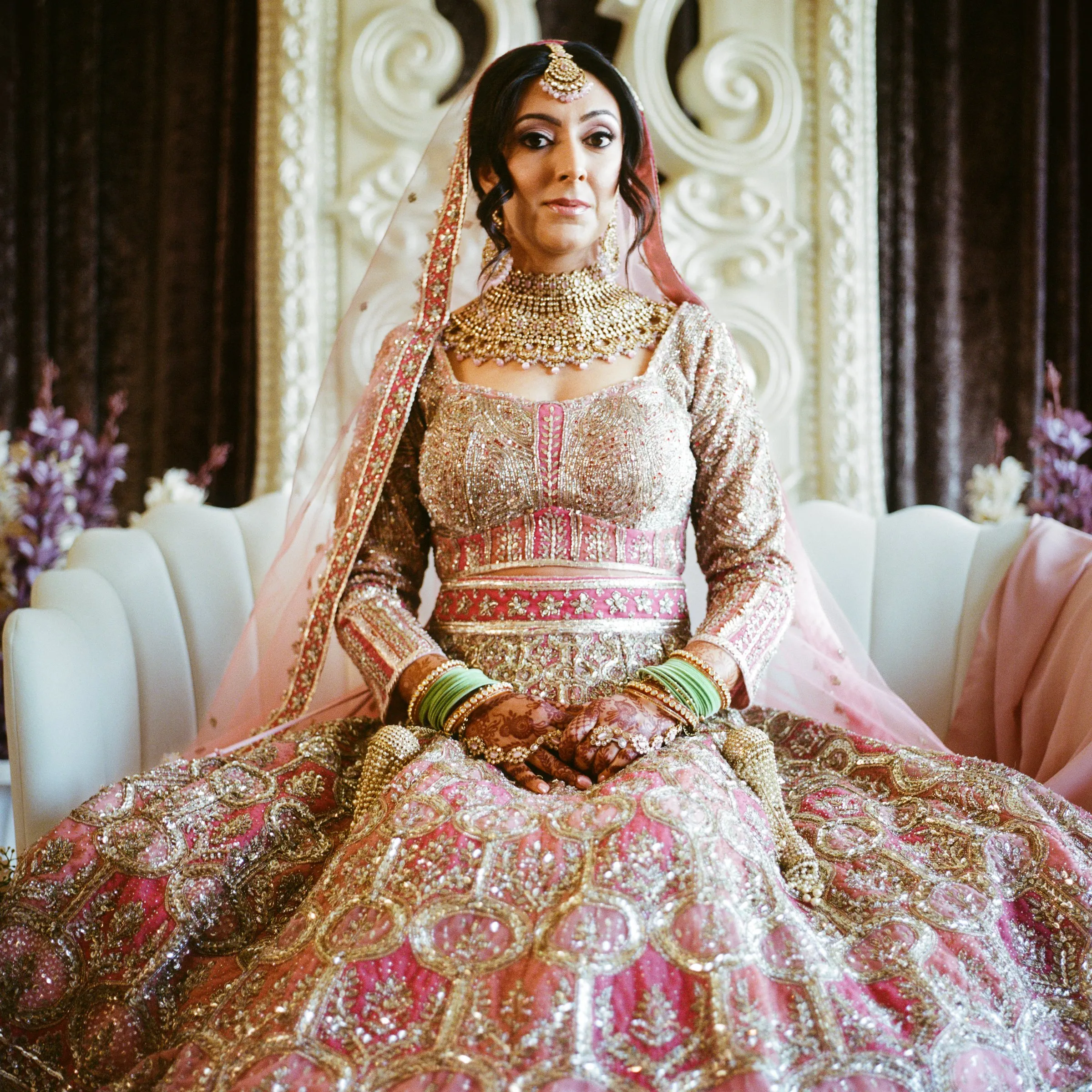 A bride in a pink embroidered lehenga sitting on a white sofa at home