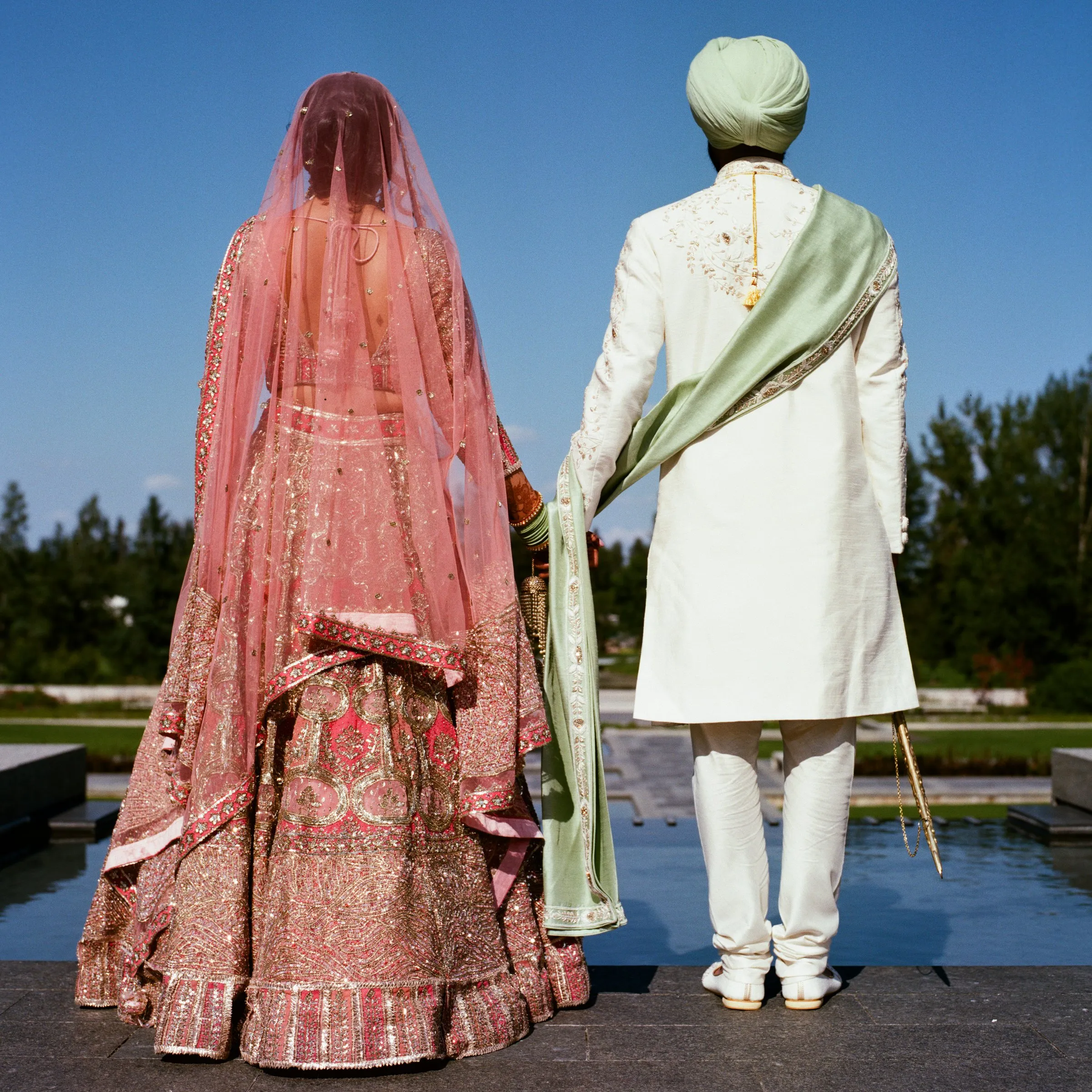 A bride and groom seen from behind beside a reflecting pool under a blue sky