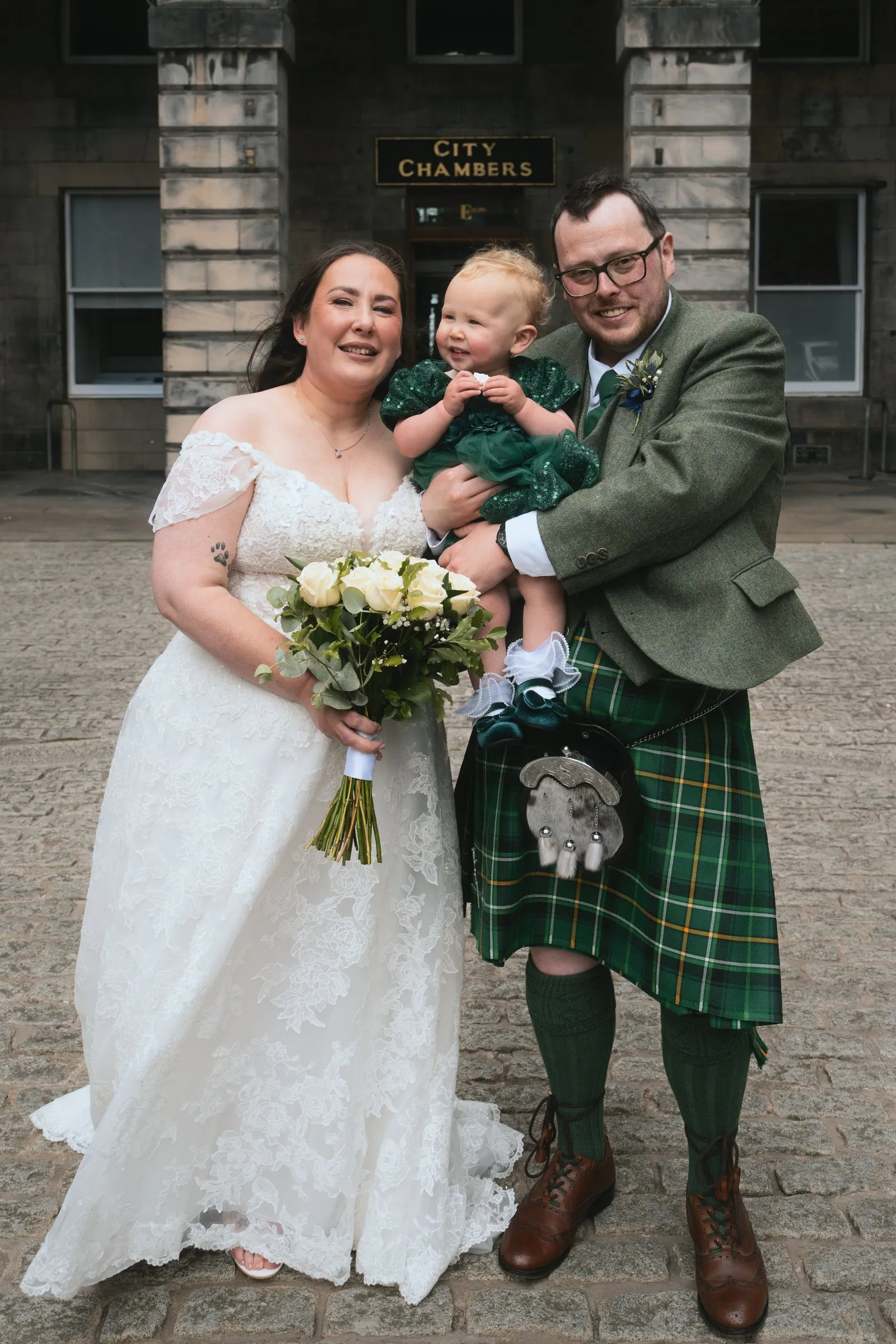 A bride and a groom in a kilt holding their baby outside the City Chambers