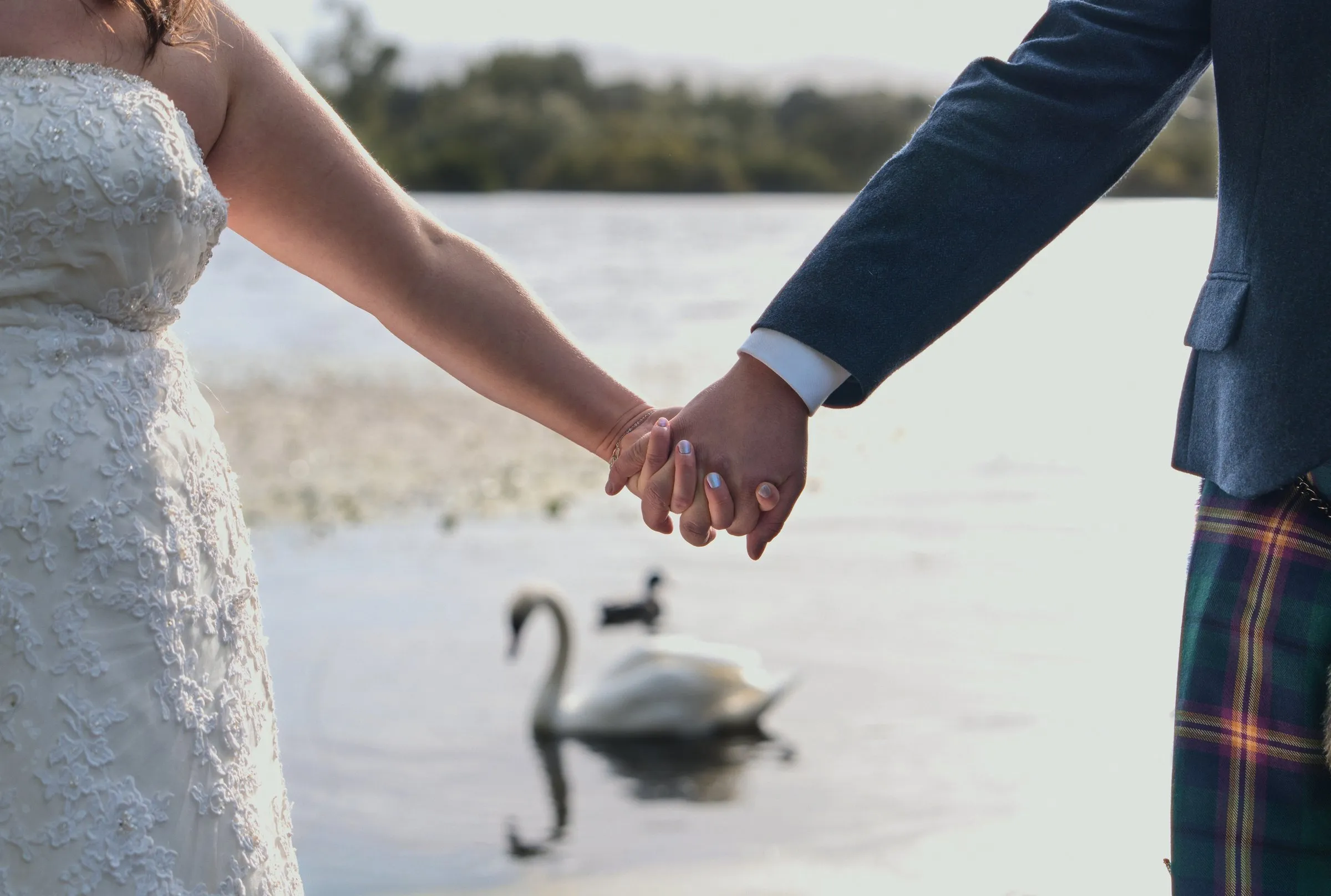 A bride and a groom in a kilt holding hands by a lake as a swan swims past