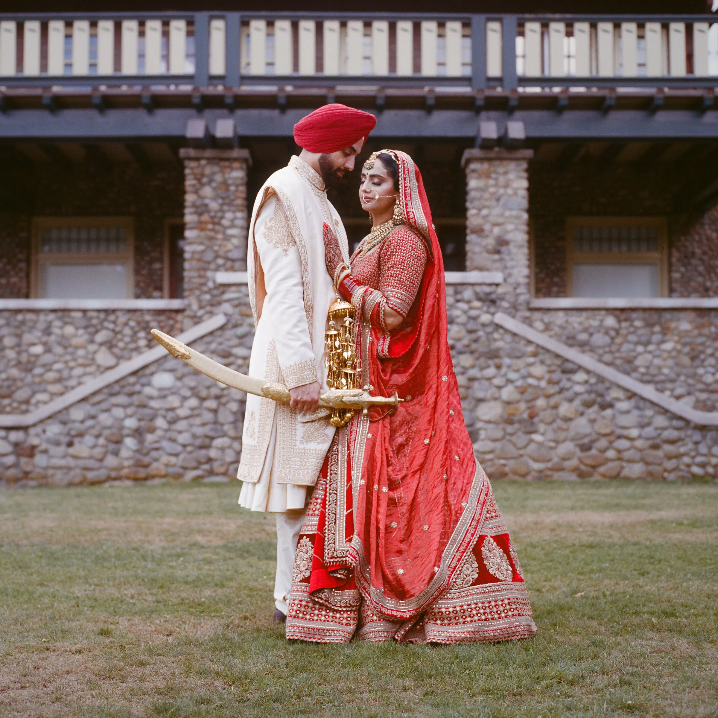 A groom in a red turban holding a sword beside his bride in front of a stone house