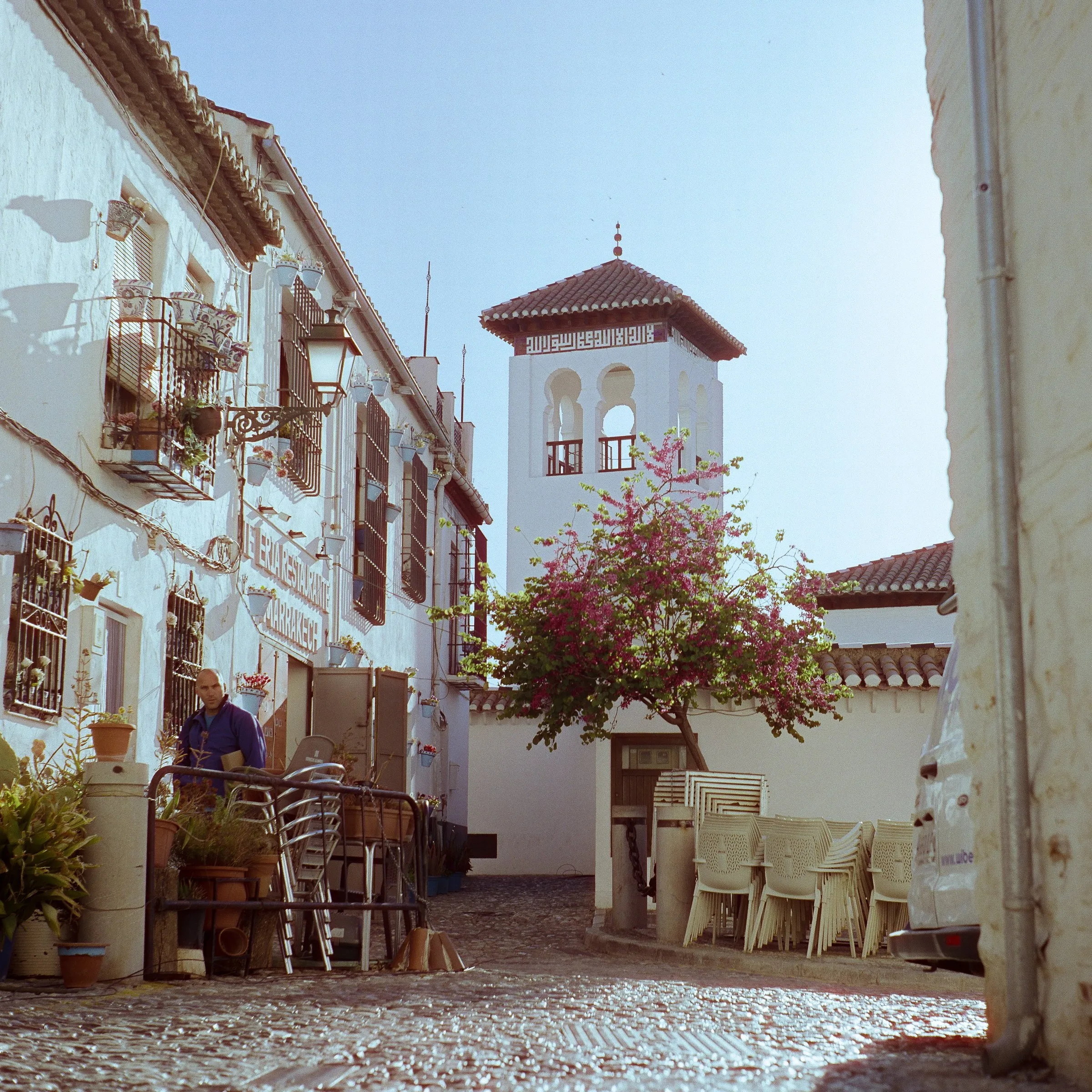 A whitewashed lane with a bell tower and pink bougainvillea, café chairs on the cobbles