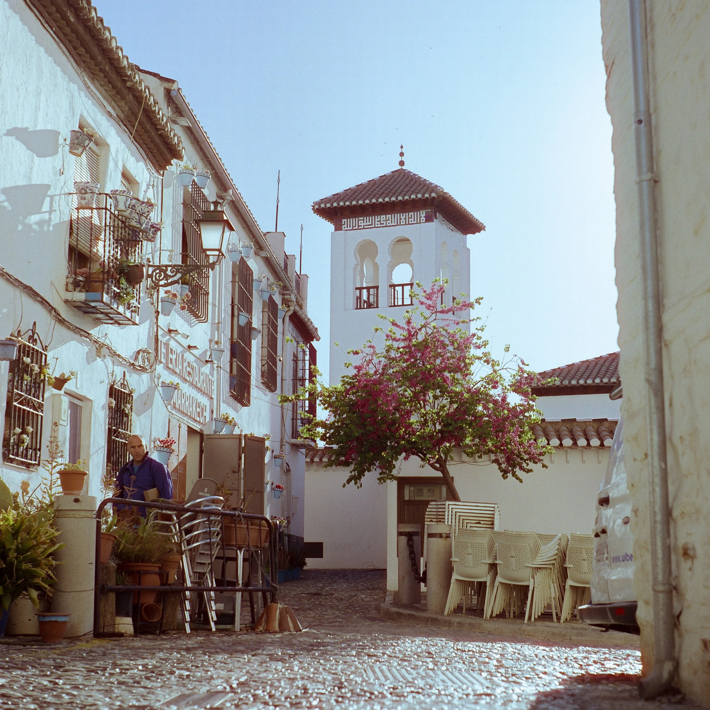 A whitewashed lane with a bell tower and pink bougainvillea, café chairs on the cobbles
