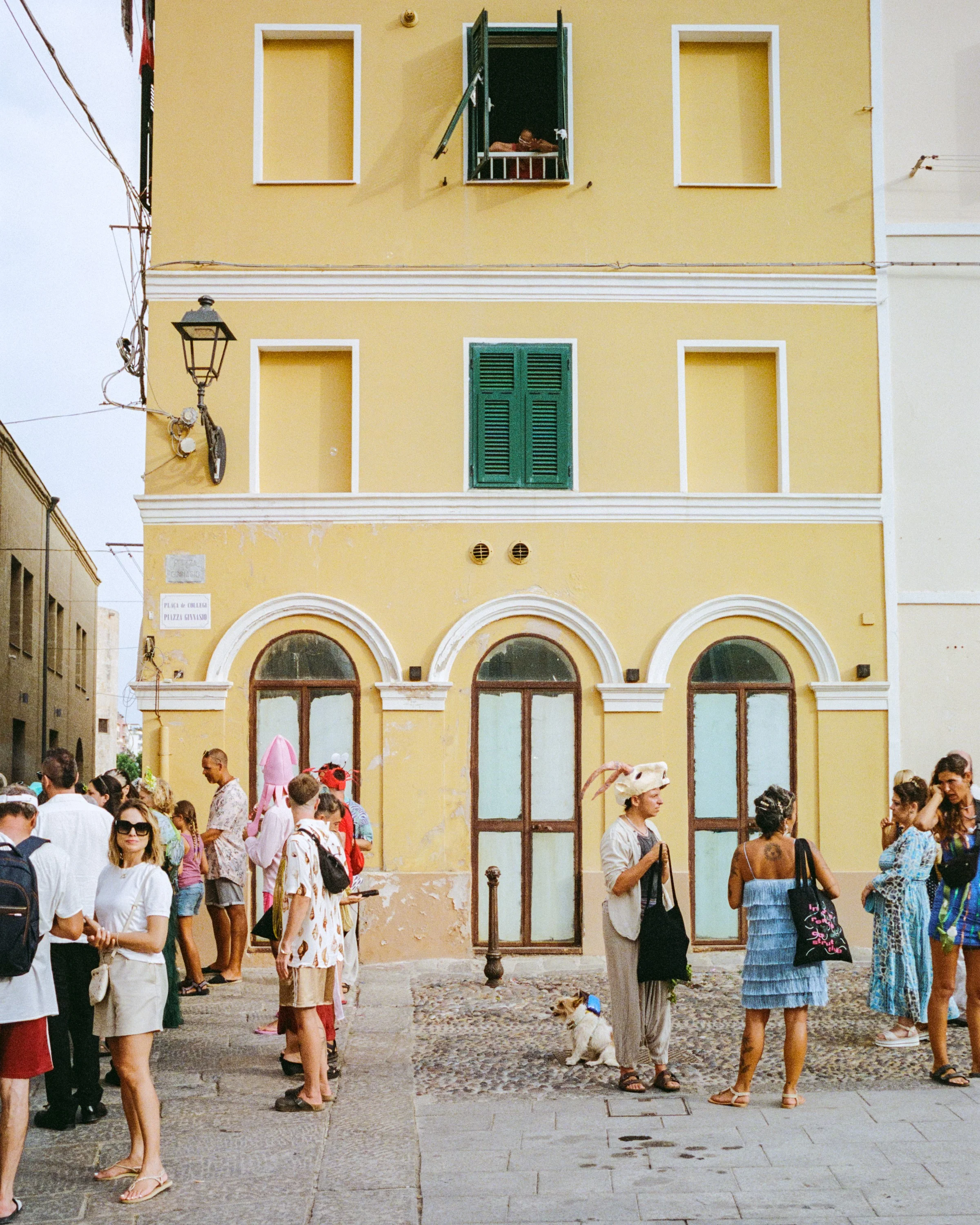 A crowd gathers in the street below a tall yellow building with arched doors